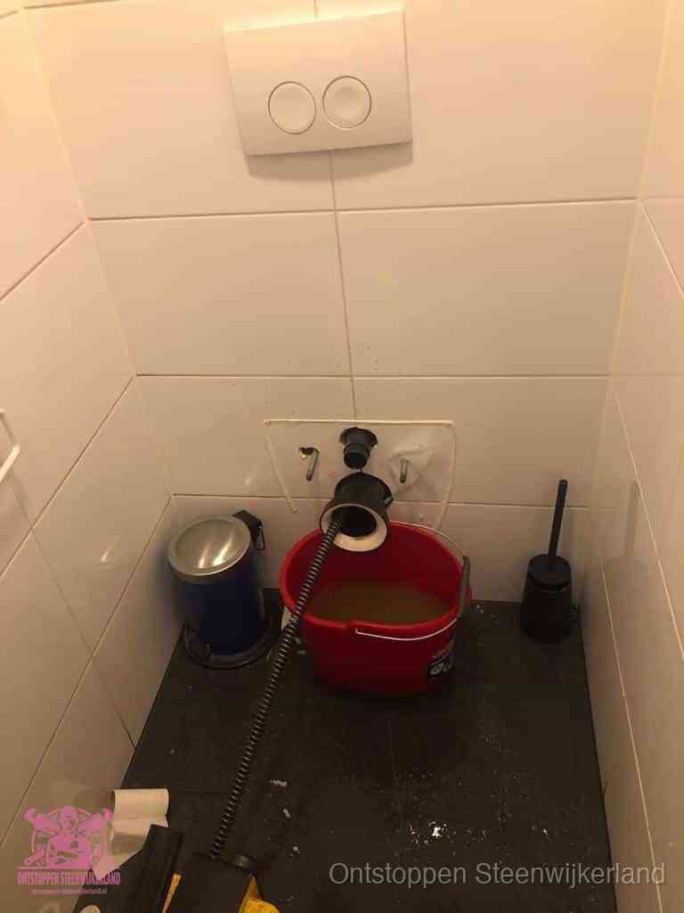 Een ontstopte wand-wc met verwijderde toiletpot waar een ontstoppingsveer in de wanduitgang verdwijnt, het achterhalen waar de verstopping zit in Steenwijkerland