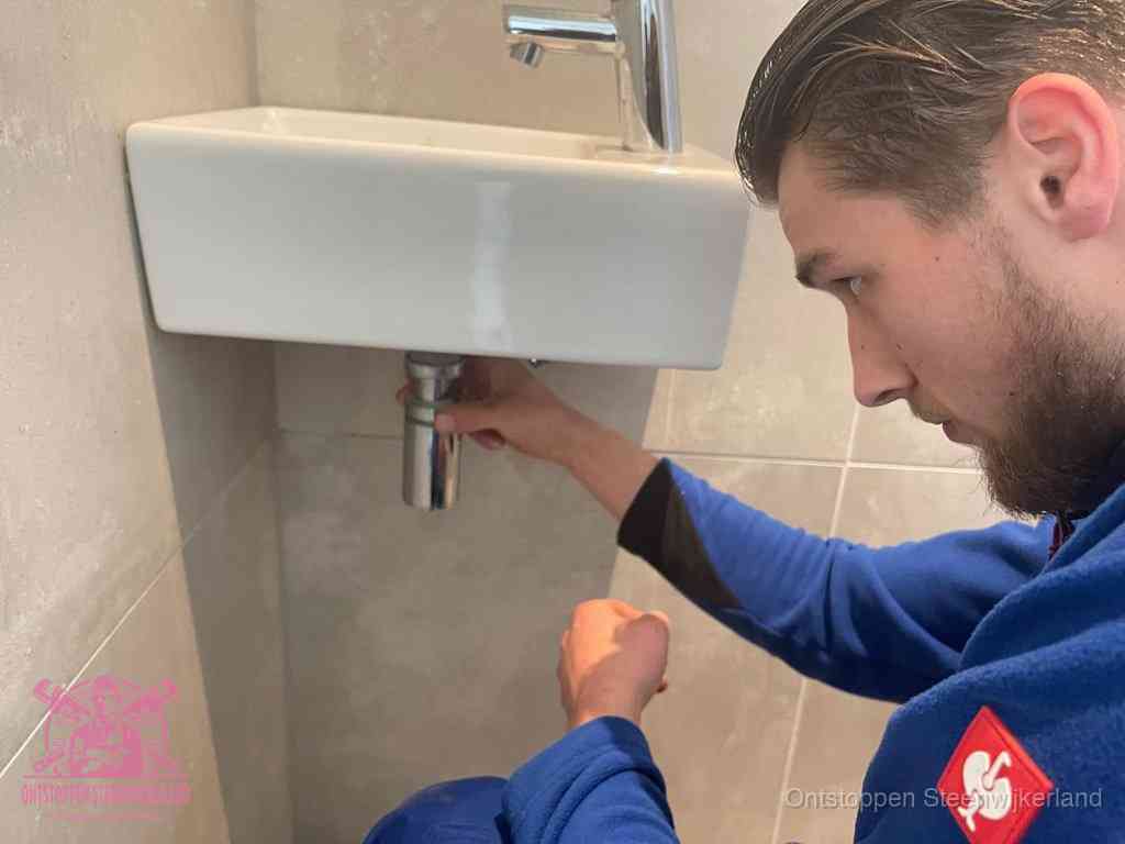 Een monteur draait met de hand de zwanenhalssifon onder een fonteinwasbak in de toiletruimte los om de bron van rioollucht in Steenwijkerland te vinden