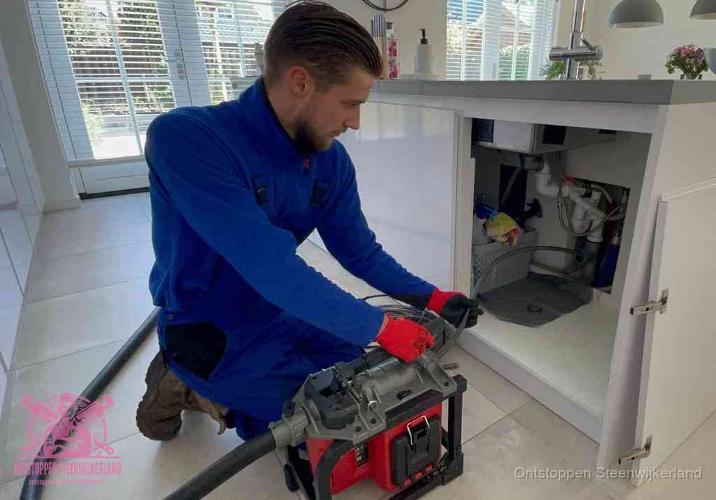 Een monteur voert een professionele elektrische ontstoppingsmachine in de afvoer onder de spoelbak tijdens het gootsteen ontstoppen in Steenwijkerland